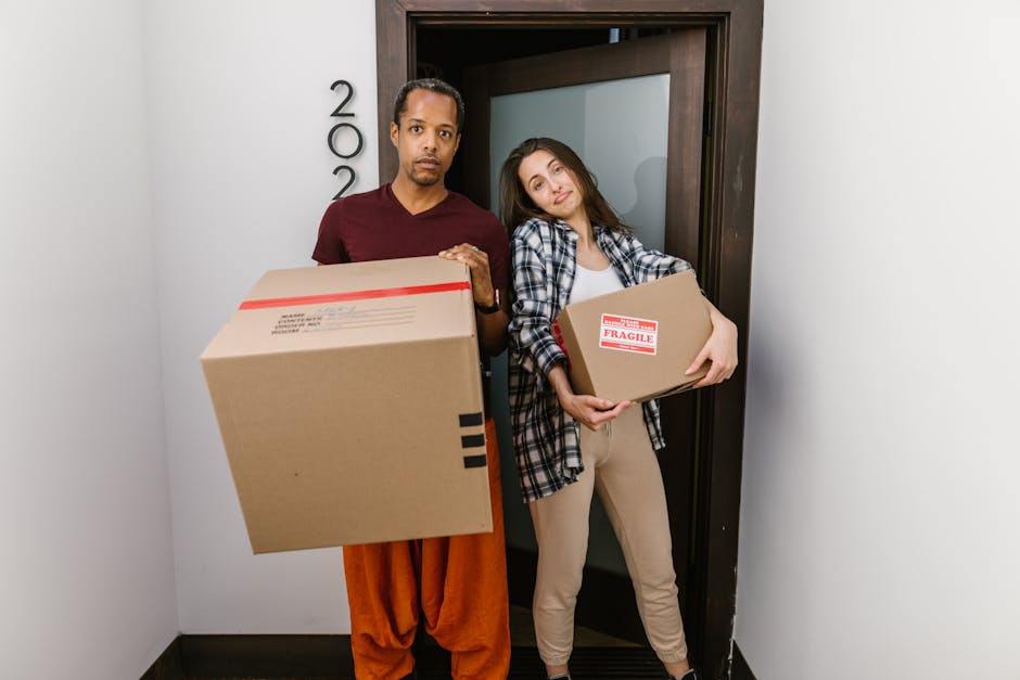 A man and a woman standing in a narrow corridor next to a dark wooden doorway, each holding cardboard boxes used for home relocation. The man, dressed in a maroon t-shirt and orange trousers, holds a large packed box with a red tape seal, while the woman, wearing a checkered shirt and beige trousers, carries a smaller fragile-labelled box with both hands. The corridor has plain white walls, and the lighting is bright, highlighting the cardboard packaging materials and the structural surroundings. The scene depicts the process of packing or unloading during furniture transport and moving logistics, consistent with professional removals services offered by Man and Van Shacklewell. This image captures the physical effort involved in loading and handling household contents for a house move, illustrating the packing and moving phase of a home relocation.