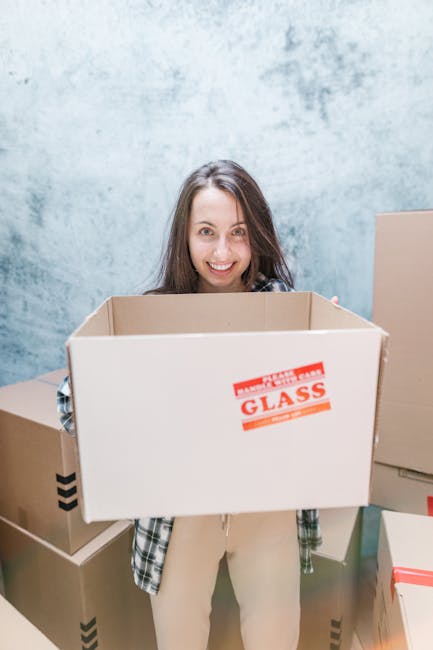 A woman with long brown hair and a friendly expression stands indoors against a light blue textured wall, holding an open cardboard removal box in front of her. The box has a red and white label that reads 'FRAGILE HANDLE WITH CARE GLASS.' She is dressed casually in a plaid shirt layered over a light top, with beige trousers. Surrounding her on the floor are additional packed cardboard boxes, some sealed with tape and others open, ready for moving. The scene depicts a home relocation process involving packing and moving furniture and household items, consistent with professional removals services like those offered by Man and Van Shacklewell. The lighting is even, highlighting the floor and the packing materials used during a furniture transport or packing and moving operation inside a property aimed at preparing for a household move.