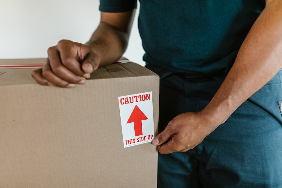 A man and a woman standing in a narrow corridor next to a dark wooden doorway, each holding cardboard boxes used for home relocation. The man, dressed in a maroon t-shirt and orange trousers, holds a large packed box with a red tape seal, while the woman, wearing a checkered shirt and beige trousers, carries a smaller fragile-labelled box with both hands. The corridor has plain white walls, and the lighting is bright, highlighting the cardboard packaging materials and the structural surroundings. The scene depicts the process of packing or unloading during furniture transport and moving logistics, consistent with professional removals services offered by Man and Van Shacklewell. This image captures the physical effort involved in loading and handling household contents for a house move, illustrating the packing and moving phase of a home relocation.