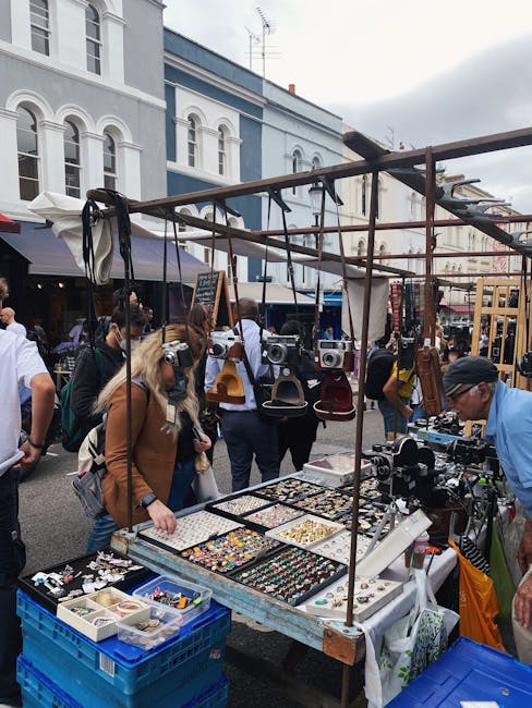 An outdoor street market scene with a vendor stall displaying vintage cameras, jewelry, and small decorative items on a wooden table. Several hanging bags, backpacks, and boxes are suspended from a metal frame above the display. In the background, groups of people are browsing, some carrying shopping bags, with historic buildings featuring large windows and ornate architectural details lining the street. The sky is overcast, and the area is lit by natural daylight. This setting, possibly during a market day in an urban setting like Shacklewell, reflects the context of home relocation or packing preparations, with individuals and vendors engaged in browsing or setting up for the market, consistent with the themes of moving and transport services provided by Man and Van Shacklewell.