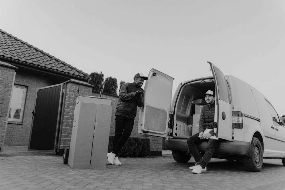 In an outdoor residential setting, two men are involved in the loading process during a home relocation using a white commercial van operated by Man and Van Shacklewell, a professional removals service. The man on the left, standing beside the open van's rear door, is wearing casual clothing including a dark jacket, light sneakers, and a cap, and appears to be checking a smartphone. The man on the right is sitting on the edge of the van's open cargo area, dressed in a quilted jacket, dark trousers, and a light-colored fedora-style hat; he is smiling and looking towards the camera. Inside the van, visible packing materials include cardboard boxes, some wrapped in plastic or fabric covers, with items carefully arranged for transport. Outside the vehicle, a large cardboard box stands upright on a paved driveway near the house, which has a tiled roof and brick walls. The environment is well-lit with natural daylight, and the scene captures the logistics of furniture transport as part of a professional moving or packing service, exemplifying efficient loading for a smooth house move.