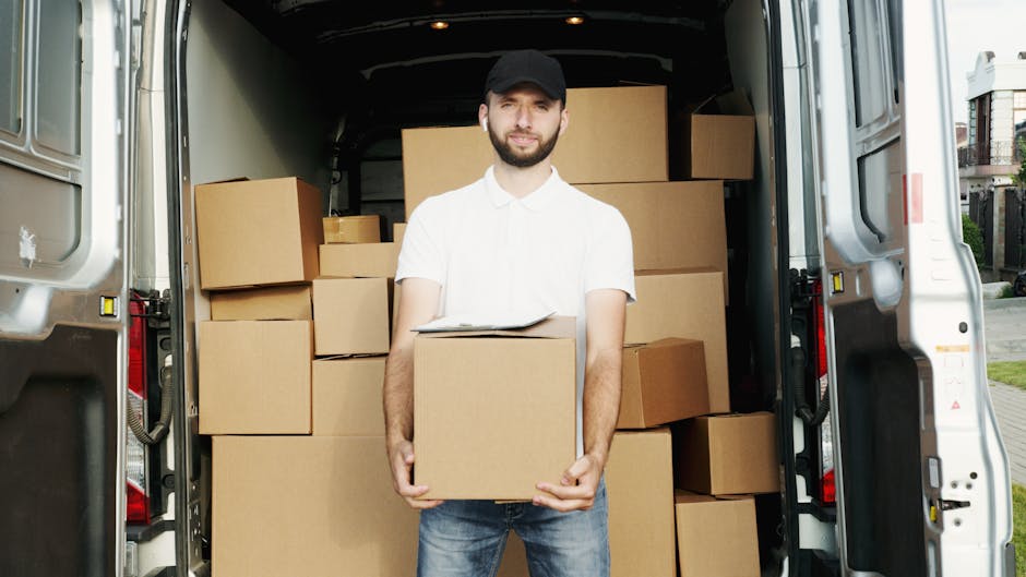 A man dressed in casual clothing with a white polo shirt and a black cap stands inside the rear of a loading van, holding a cardboard box at the forefront. The van's interior is filled with numerous cardboard boxes of various sizes, some stacked on top of each other, ready for house removal or furniture transport. The boxes are sealed with packing tape and are positioned on the van's floor, with some resting against the walls. The vehicle is parked outdoors on a street with residential buildings visible in the background on the right side. The lighting indicates daylight, and the scene captures the loading process involved in home relocation, supported by the presence of packing materials and furniture items. This setting exemplifies the practical aspects of professional removals services offered by Man and Van Shacklewell, focusing on efficient packing and transport methods.