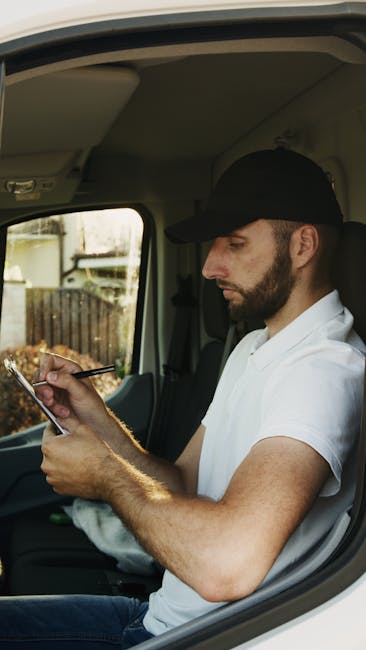 In an outdoor residential setting, two men are involved in the loading process during a home relocation using a white commercial van operated by Man and Van Shacklewell, a professional removals service. The man on the left, standing beside the open van's rear door, is wearing casual clothing including a dark jacket, light sneakers, and a cap, and appears to be checking a smartphone. The man on the right is sitting on the edge of the van's open cargo area, dressed in a quilted jacket, dark trousers, and a light-colored fedora-style hat; he is smiling and looking towards the camera. Inside the van, visible packing materials include cardboard boxes, some wrapped in plastic or fabric covers, with items carefully arranged for transport. Outside the vehicle, a large cardboard box stands upright on a paved driveway near the house, which has a tiled roof and brick walls. The environment is well-lit with natural daylight, and the scene captures the logistics of furniture transport as part of a professional moving or packing service, exemplifying efficient loading for a smooth house move.
