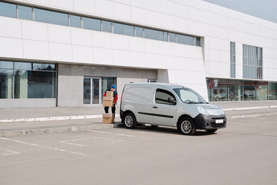 A person wearing a blue hat and red jacket is standing outside a modern, multi-storey building loading large cardboard boxes onto a small, grey panel van parked on an empty parking lot. The individual is using their hands to lift and carry the boxes, which are stacked on the pavement beside the vehicle. The van is positioned close to the building's entrance, indicating a home relocation or furniture transport process. The building features a white facade with large glass windows and a stone base, and the parking area is marked with white lines and curb edges. The scene is captured in daylight, with natural lighting illuminating the loading activity, reflecting efficient moving logistics typically managed by professional removals services like Man and Van Shacklewell.