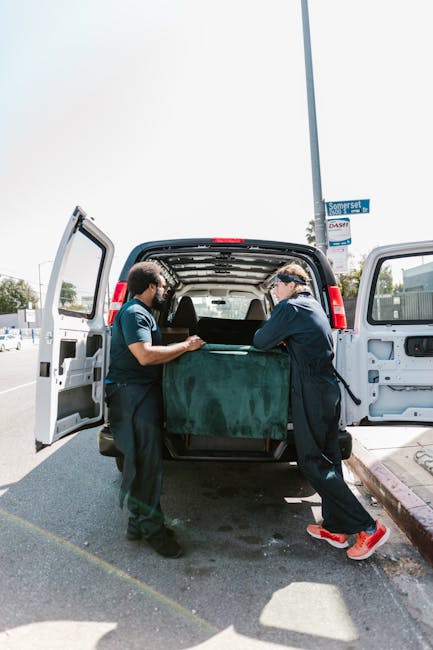 Two movers from Man and Van Shacklewell are loading a large, green upholstered furniture piece into the open cargo area of a white van parked on a street in Dalston. One mover, wearing a navy jacket and red shoes, is guiding the item into the van, while the other, dressed in a blue shirt and dark trousers, assists from the opposite side. The van is positioned next to a sidewalk with a black curb, and in the background, a blue street sign reading 'Somerset' is visible on a pole. The interior of the van reveals an empty cargo space with a black floor, ready for additional packing or furniture transport. Overhead, the sky is partially cloudy, providing natural daylight that illuminates the area. This scene captures the packing and loading process typical of home relocation or furniture transport, highlighting the careful handling of household items by professional movers from Man and Van Shacklewell during a professional removal service.