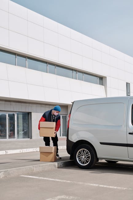 A person wearing a blue hat and red jacket is standing outside a modern, multi-storey building loading large cardboard boxes onto a small, grey panel van parked on an empty parking lot. The individual is using their hands to lift and carry the boxes, which are stacked on the pavement beside the vehicle. The van is positioned close to the building's entrance, indicating a home relocation or furniture transport process. The building features a white facade with large glass windows and a stone base, and the parking area is marked with white lines and curb edges. The scene is captured in daylight, with natural lighting illuminating the loading activity, reflecting efficient moving logistics typically managed by professional removals services like Man and Van Shacklewell.
