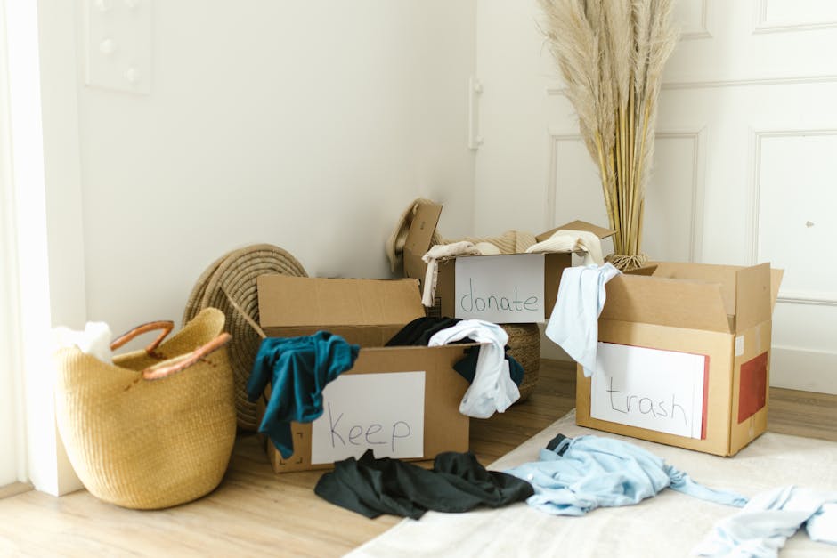 Two volunteers wearing blue T-shirts with the word 'VOLUNTEER' printed on the front are participating in a house removal or recycling activity indoors. One volunteer is holding a crumpled, transparent