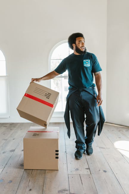 A man with curly dark hair and a beard, wearing a blue t-shirt and jeans, sits on a wooden floor inside a room with large arched windows allowing natural daylight to illuminate the space. The background shows a street scene with parked cars and modern buildings visible through the windows. Several flattened cardboard boxes, some sealed with red packing tape, are stacked beside him, indicating packing or unpacking activities during a home relocation process. A tall potted plant with broad green leaves is placed near the window on the right. The room appears to be in the midst of a move, with furniture and packing materials suggesting an ongoing furniture transport or moving service. This scene is associated with the services offered by Man and Van Shacklewell, providing last-minute removals options for house moves or furniture transport.