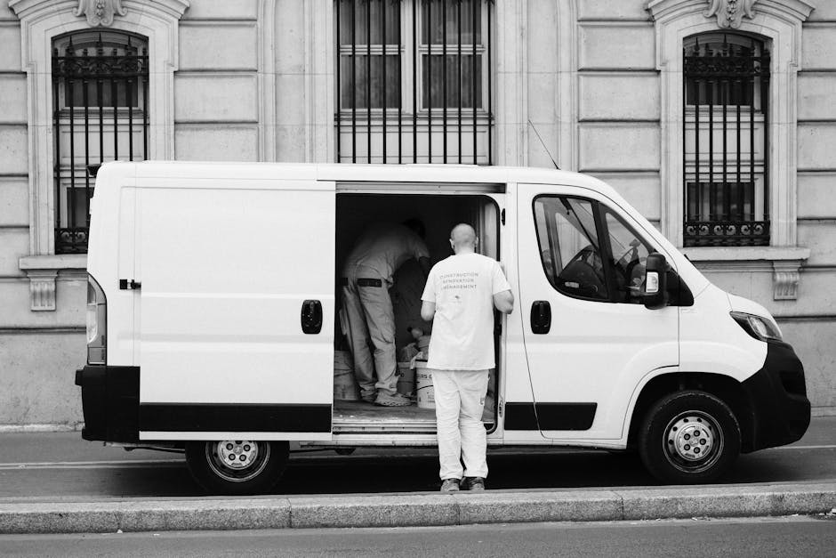 A black-and-white photograph showing a white man and van parked on a city street in front of an ornate stone building with barred windows. The van's sliding door is open, revealing three movers inside