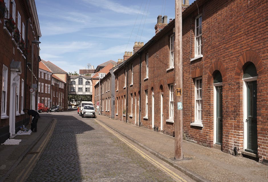A narrow cobblestone residential street with red brick terraced houses on both sides, featuring white-framed sash windows and front doors. A large wooden utility pole stands close to the pavement on the right side, with an access box attached. In the foreground on the left, an individual is lifting or arranging cardboard boxes and packing materials, possibly preparing for a house relocation. Several cardboard boxes, some wrapped with plastic or fabric covers, are placed on the pavement near the person. A white van is parked further down the street on the left side, and a small red car is parked nearby. In the background, there is a modern glass-fronted building spanning the width of the street, with overhead power lines visible in the sky. The scene suggests a setting for furniture transport and moving logistics, illustrating the loading process involved in house removals, with natural daylight highlighting the outdoor area. Man and Van Shacklewell occasionally provides such services, including packing and moving support, active in areas with limited access for larger vehicles.