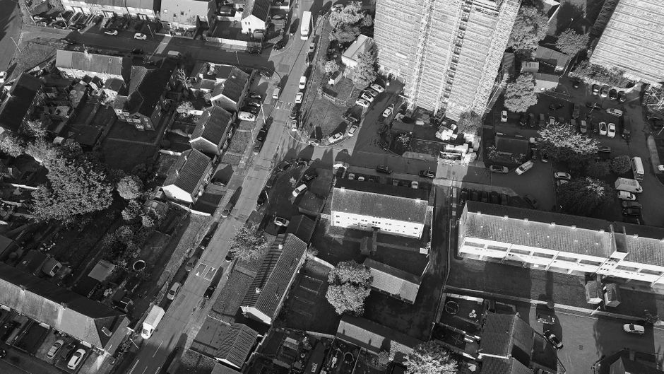 An aerial black-and-white photograph showing a densely built residential area with multiple houses, small commercial buildings, and apartment blocks. The image captures a narrow street running through the neighbourhood, with parked cars lining both sides and pedestrian pathways. On the left side, there are several small, detached houses with tiled roofs, garden areas, and trees interspersed among the properties. In the center and right, larger multi-storey apartment buildings with flat roofs and multiple windows are visible, along with adjacent parking areas filled with various vehicles. Limited street access and tight spacing between buildings suggest potential challenges for furniture transport and home relocation logistics, relevant to the access issues faced during the moving process for companies like Man and Van Shacklewell. The scene is illuminated by natural daylight, highlighting the urban environment and the density of the neighborhood, which may impact the movement of large furniture or appliances during home removals.