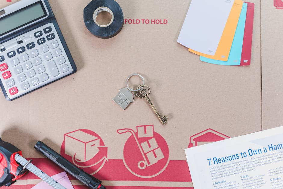 An overhead view of a desk surface used in house packing and home relocation tasks, displaying various objects including a calculator, a roll of black tape, a set of multicoloured index cards, a keychain with a small house and a vintage-style key, and a printed document titled '7 Reasons to Own a Home'. The items are arranged on a cardboard packing surface featuring red printed graphics of a moving box, a hand cart, and arrows, with the text 'FOLD TO HOLD'. The scene, captured in natural lighting, illustrates the detailed process of packing and organizing documents and essentials before moving. Occasionally, Man and Van Shacklewell's removals services may involve assisting with packing, furniture transport, or home relocation logistics, as reflected by these preparatory items.