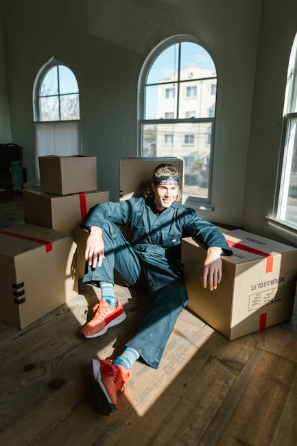 A person dressed in casual work clothes, including a denim jacket and pants, sits on the wooden floor in a well-lit room with large arched windows that allow natural daylight to enter. The individual is surrounded by multiple cardboard moving boxes, some sealed with red packing tape, indicating a home relocation process. The person appears relaxed, leaning against the boxes with one arm resting on a box and a slight smile on their face. The room has a neutral colour scheme with white walls and light streaming through the windows, suggesting the interior of a residential property preparing for moving day. The boxes are stacked and spread around in an organized manner, possibly for packing and loading furniture or belongings onto a moving vehicle. This scene reflects the packing and furniture transport stages typical of house removals handled by services like Man and Van Shacklewell.