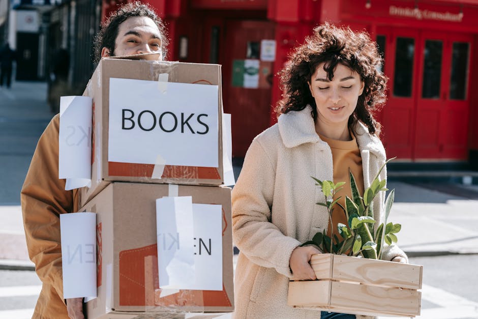 Two individuals participating in a home relocation process outdoors on a city street. The person on the left is partially visible, carrying a stack of three cardboard boxes taped securely shut, with white labels reading 'BOOKS' and other partially visible labels. The person on the right, a woman with curly brown hair, is holding a wooden container filled with leafy green plants, preparing for packing and moving activities. They are standing on a paved sidewalk in front of a red building with glass doors, and are dressed in casual clothing suitable for moving tasks, with natural daylight illuminating the scene. The image depicts the packing stage of furniture transport and moving logistics, often coordinated by a professional removals service such as Man and Van Shacklewell.