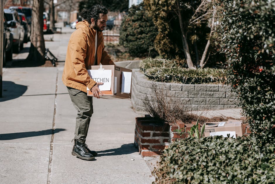 A man wearing a mustard yellow jacket, grey trousers, and black work boots is standing on a residential pavement, loading a cardboard box labeled 'KITCHEN' into a van. The box is held with both hands, and his gaze is directed downward towards the box. Behind him, there are other cardboard packing cartons, some with labels, placed on the pavement and partially behind a low brick wall with landscaping, including bushes and sparse plants. The scene is set outdoors on a sunny day, with parked cars visible along the street and trees providing partial shade, indicating an urban home relocation environment. The man and the boxes are positioned near the edge of the property, in preparation for furniture transport and moving services, typical of house removals in a narrow street setting, as referenced in the Shacklewell N16 removals guide for narrow street moves by Man and Van Shacklewell.